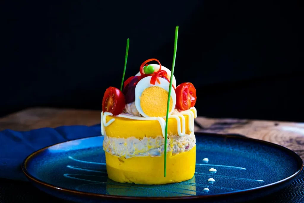 Peruvian Cuisine Guide: The Most Iconic Dishes and Drinks You Must Try 7 Traditional Causa Limeña layered with mashed yellow potatoes, avocado, chicken, and boiled eggs, beautifully presented with ají amarillo sauce.